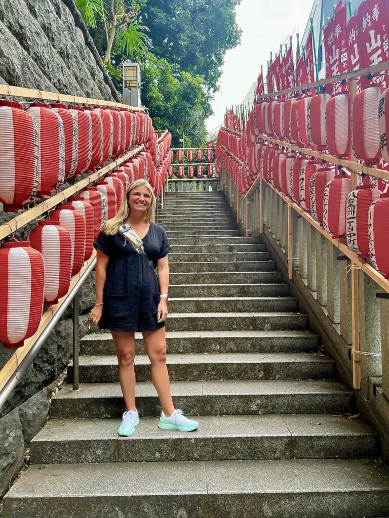 Smiling person in a black outfit stands on stone steps lined with red and white lanterns. Lush greenery is visible in the background.