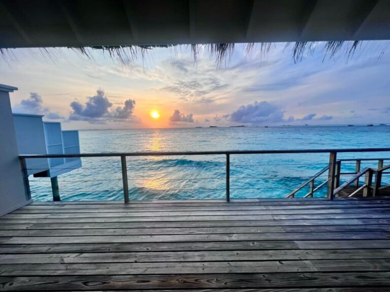 Wooden deck overlooking calm ocean waters at sunset. The sun casts a warm glow, with orange and blue hues blending in the sky. Peaceful atmosphere.
