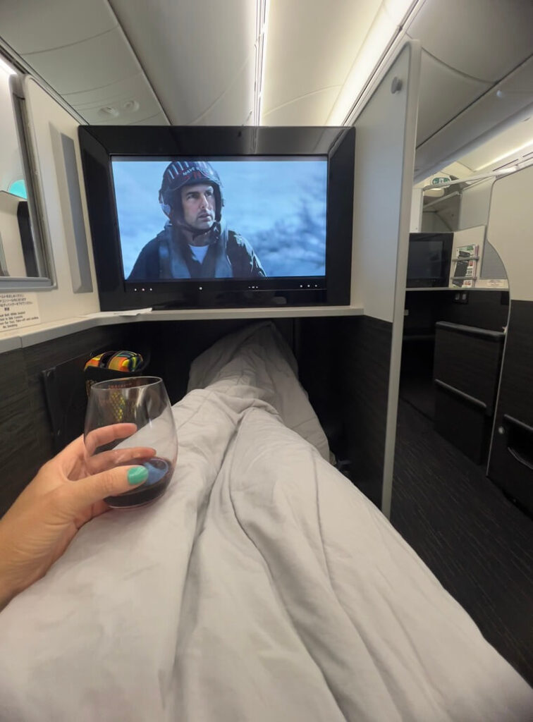 A person in an airplane's business-class cabin relaxes under a white blanket, watching a movie on a screen while holding a glass of red wine.