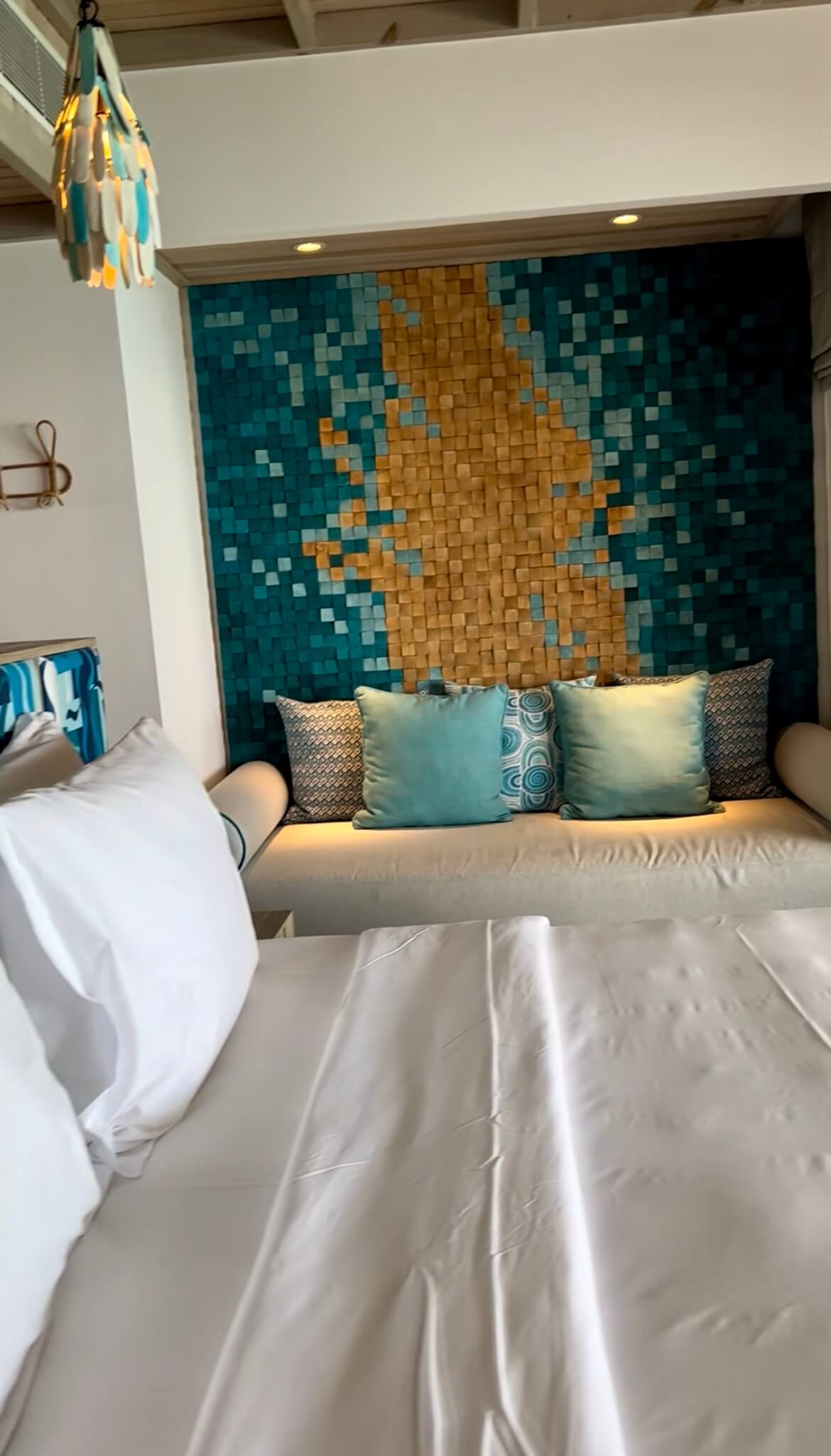 Calming bedroom with a neatly made bed, white sheets, and decorative pillows. A textured mosaic wall in teal and beige adds artistic flair.