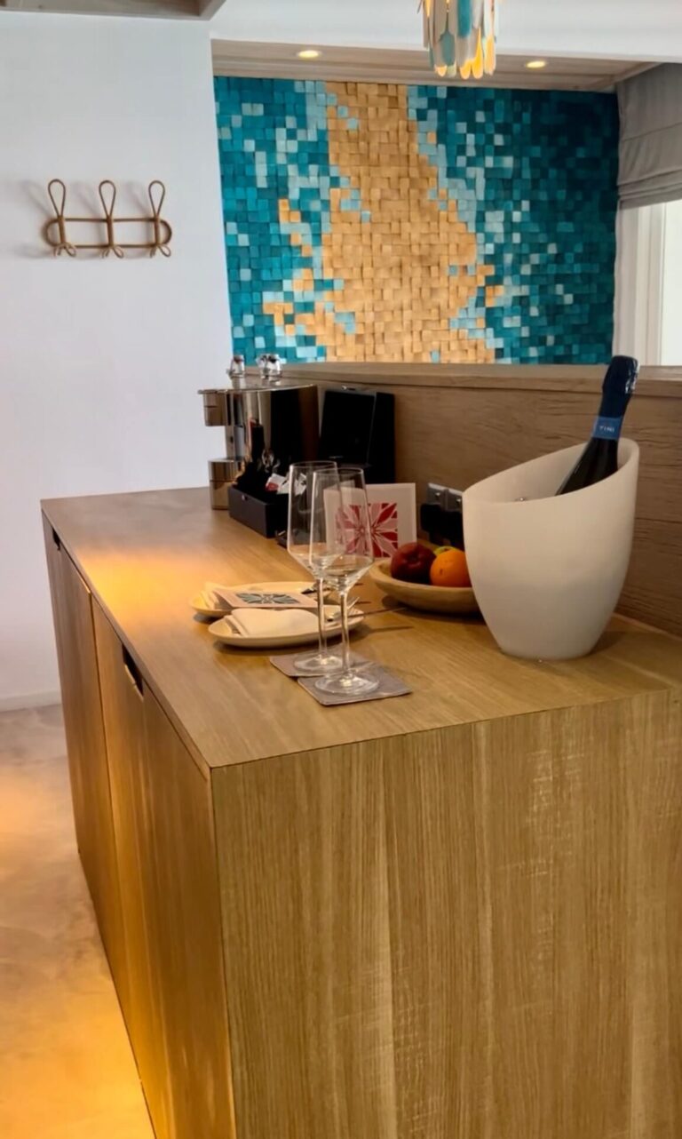Modern kitchen counter with wooden design, featuring two wine glasses, a white wine cooler, a fruit bowl, and a colorful mosaic wall backdrop.