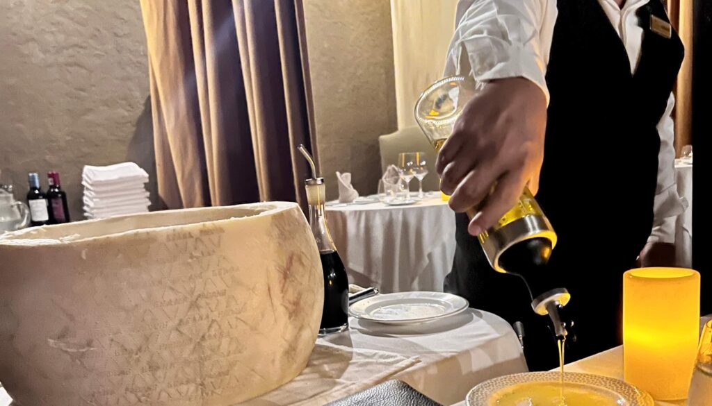 A server in a white shirt and black vest is pouring olive oil onto a plate. A large wheel of cheese, bottles, and folded napkins are on the table. The setting is a softly lit restaurant, conveying a refined and elegant atmosphere.