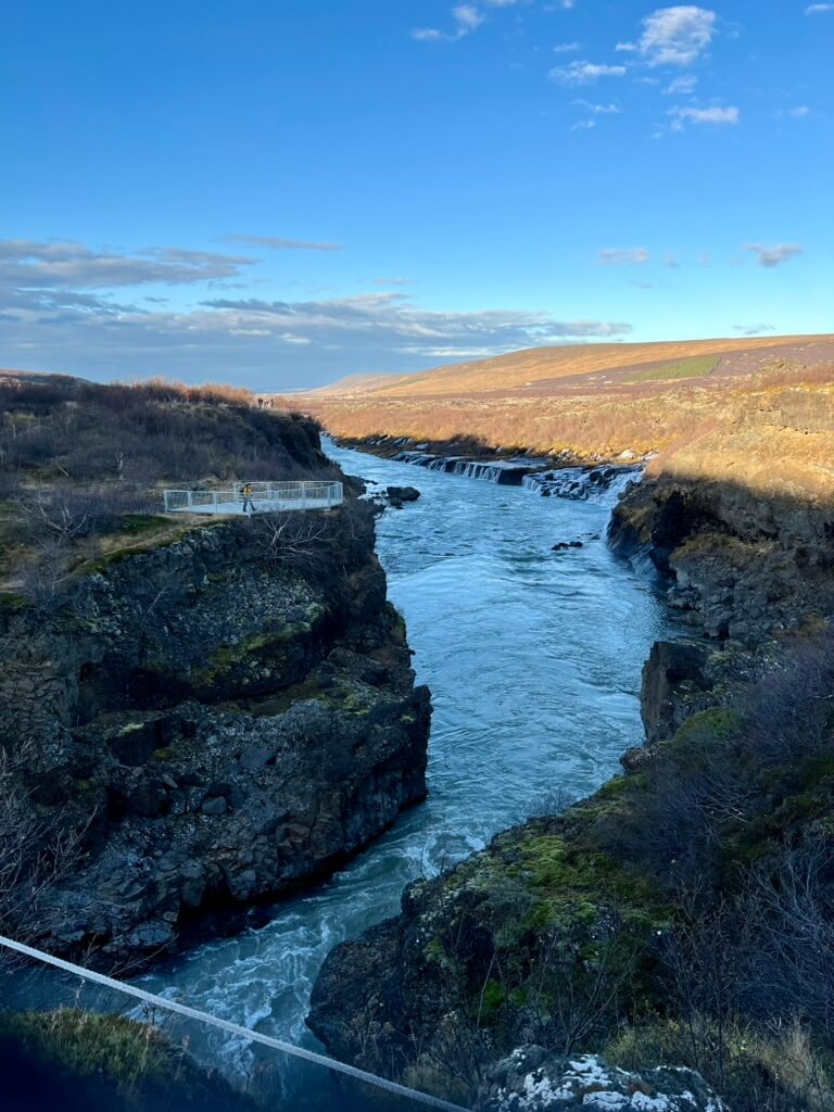 A narrow river flows briskly through a rocky gorge under a clear blue sky. Sparse vegetation and a viewing platform are visible, suggesting a serene, natural landscape.
