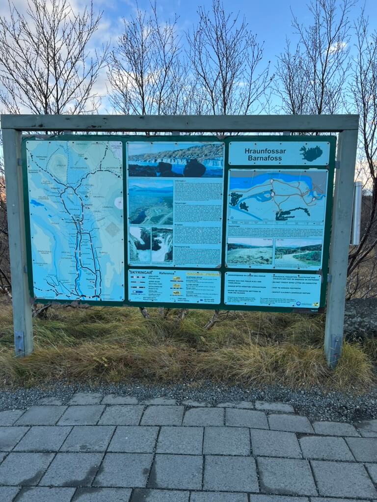Informational sign for Hraunfossar and Barnafoss waterfalls with maps and text, set against bare winter trees on a stone path. Calm, educational tone.