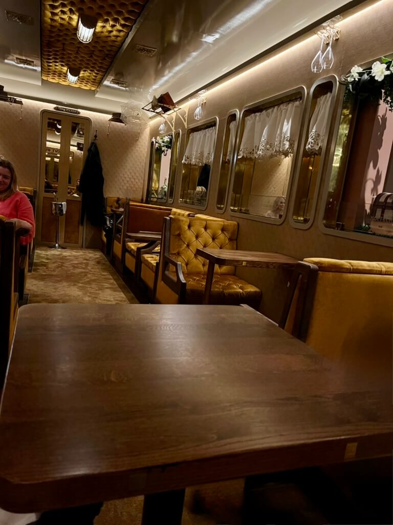 Luxurious vintage train interior with tufted leather seats, wooden tables, ornate ceiling, and soft lighting. A person in a pink sweater sits on the left.