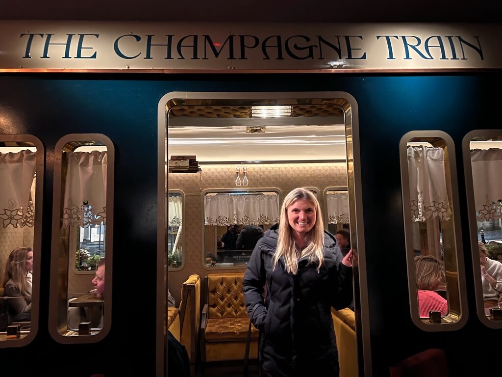 A woman in a black coat smiles at the entrance of "The Champagne Train," with an elegant interior featuring golden seats and decorative curtains.