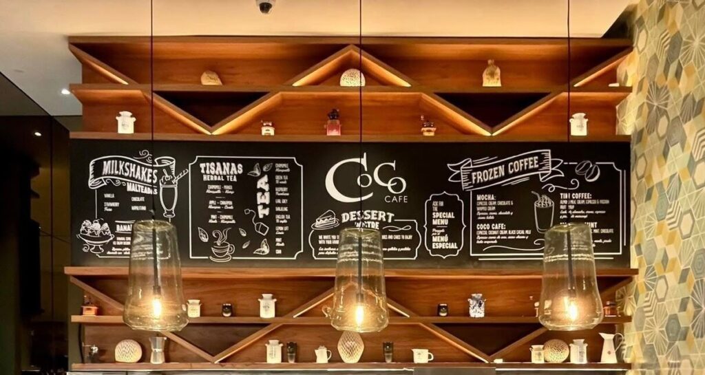 Chic café interior with wooden shelves and pendant lights. A blackboard menu lists milkshakes, teas, and frozen coffee. Warm and inviting ambiance.