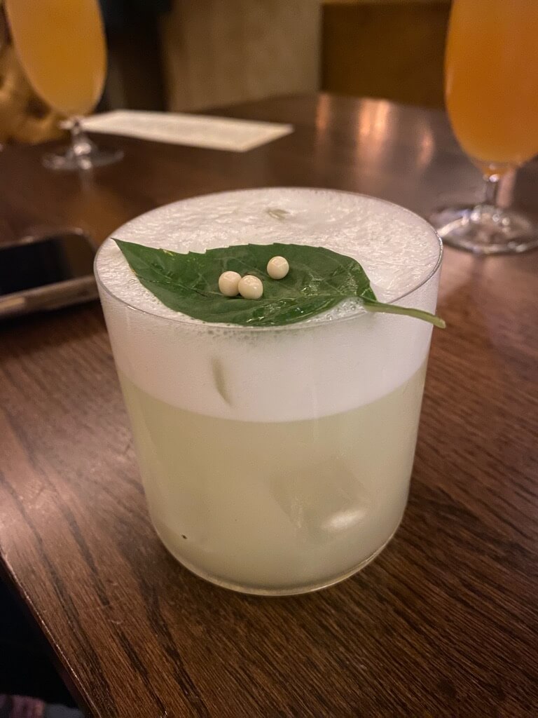 A cocktail glass sits on a wooden table, filled with a frothy light green drink. A green leaf with three small white spheres garnishes the top.