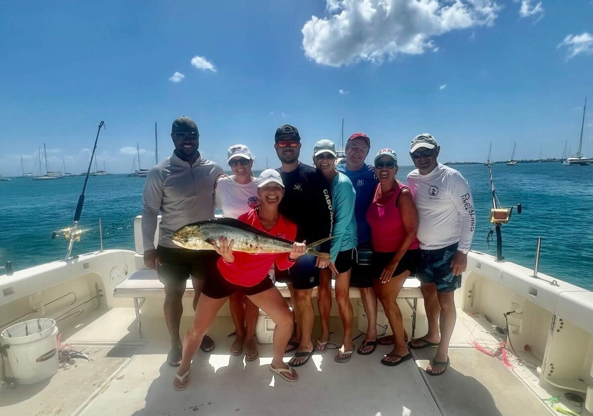 A group of eight people smiling on a boat under a clear sky, with one person holding a large fish. The ocean is calm, and sailboats are visible in the background.