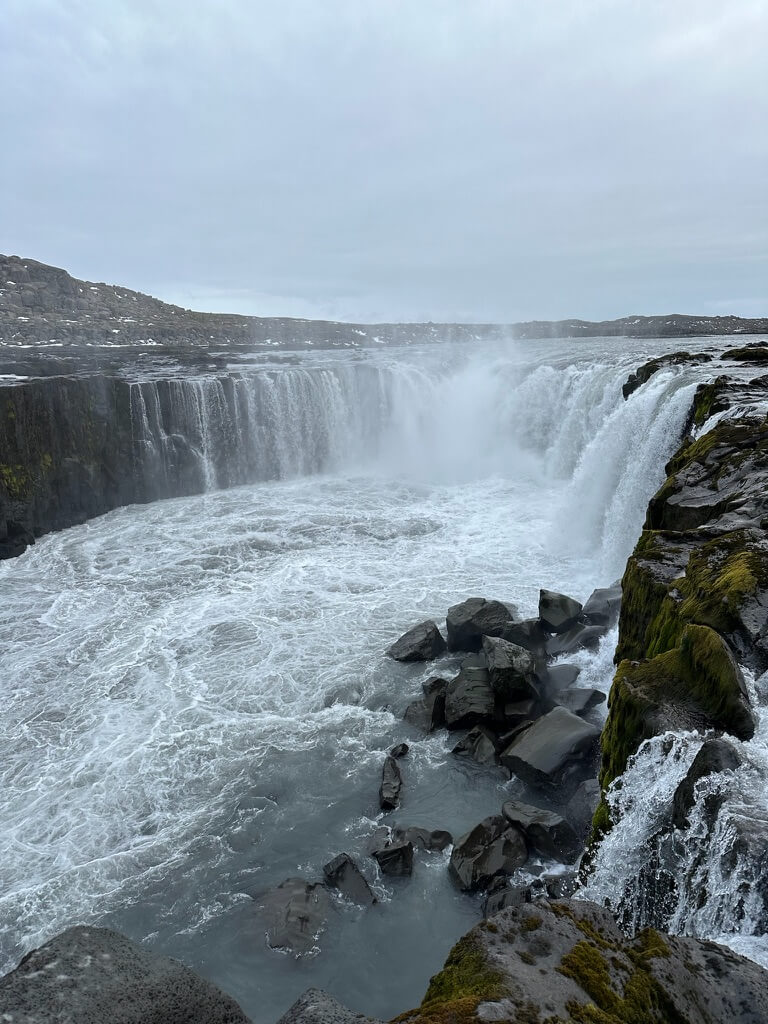Powerful waterfall cascading over rocky cliffs into a turbulent pool. Moss-covered rocks border the scene under an overcast sky, evoking a serene yet dramatic mood.