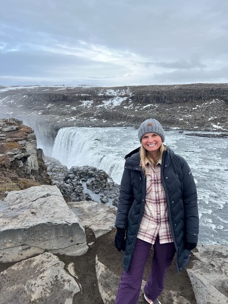 A woman in a gray beanie and dark coat smiles near a large, thundering waterfall with rocky cliffs and patches of snow under a cloudy sky.