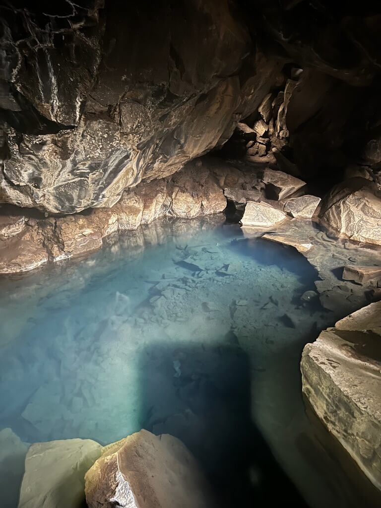Tranquil cave with clear blue water reflecting rugged stone walls, casting serene shadows. The atmosphere is peaceful and mysterious.
