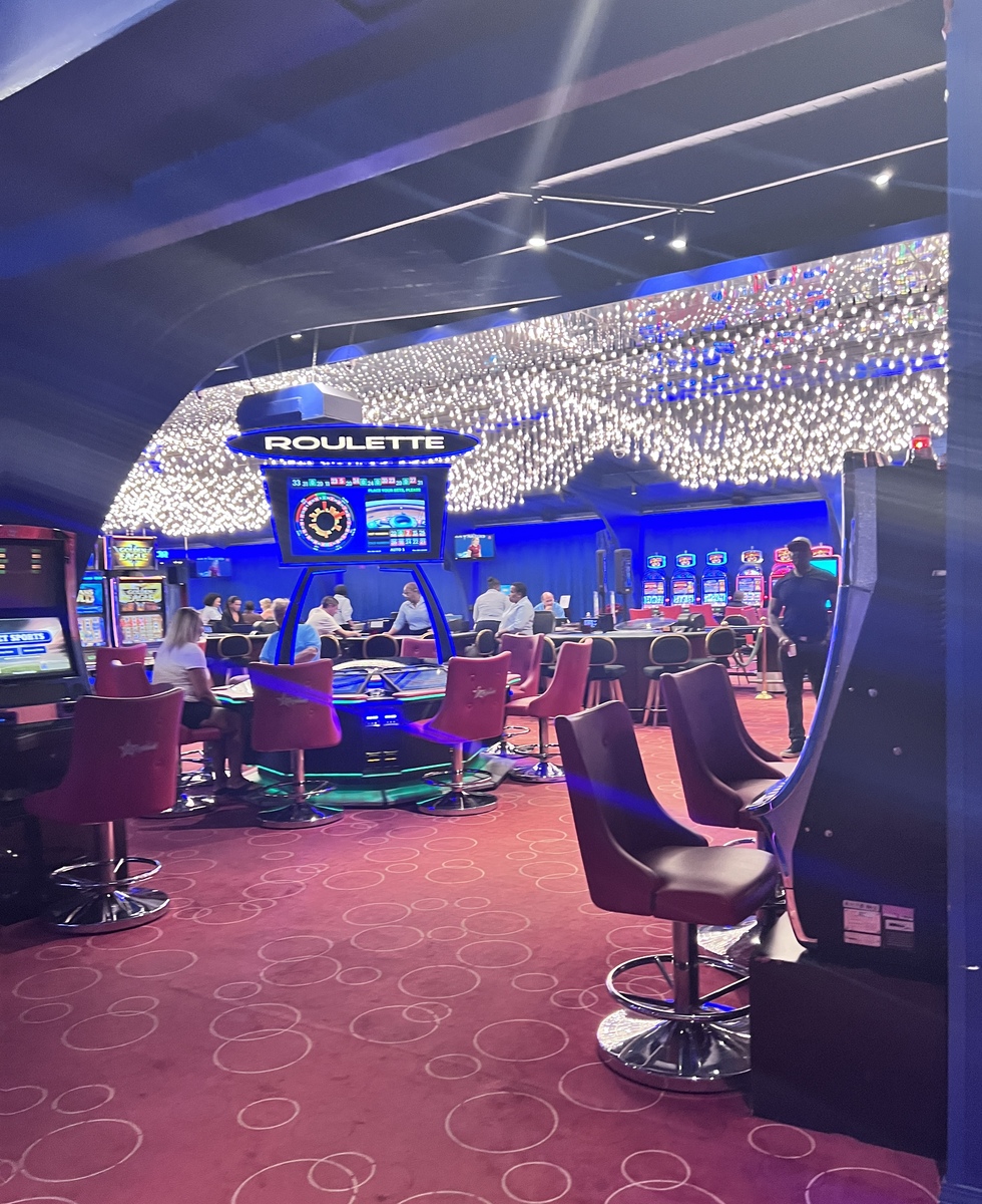 A vibrant casino scene with people gathered around a roulette table under dazzling, star-like ceiling lights. Slot machines line the back, creating an energetic atmosphere.