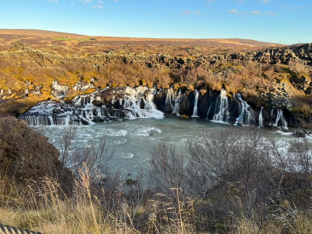 Rocky waterfall cascades into a river surrounded by rugged landscape. Bare trees and grasses in the foreground; vast, barren horizon under a clear blue sky.