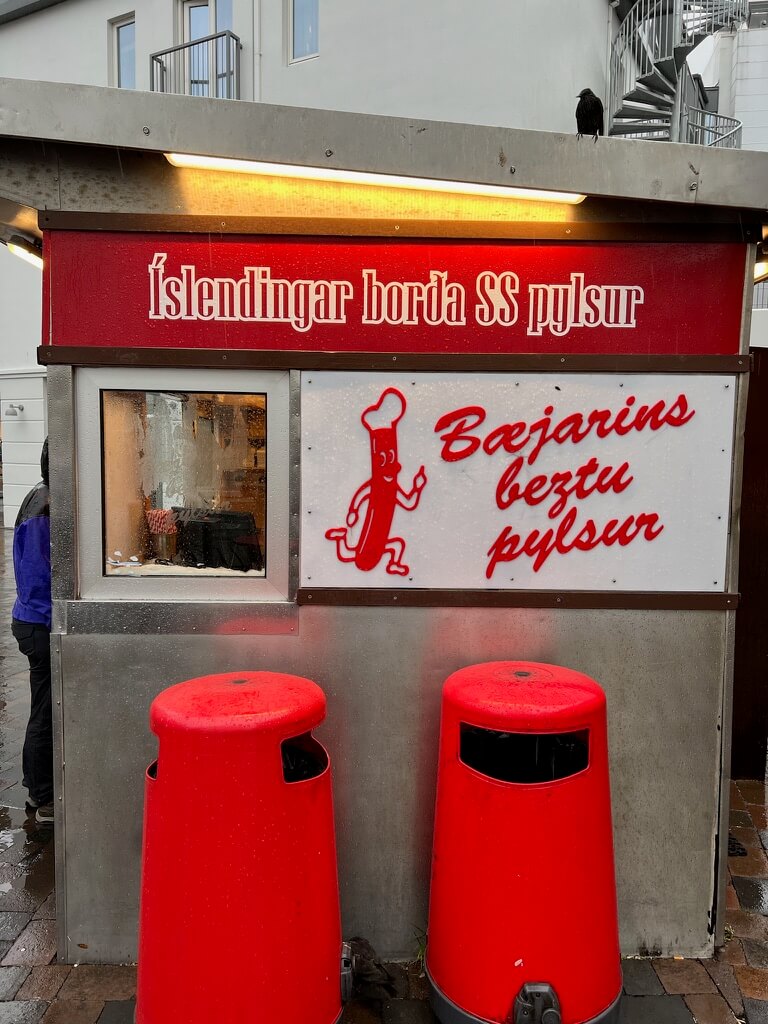 A small outdoor hot dog stand with a red sign reading “Íslendingar borða SS pylsur.” Two bright red trash cans sit in front, and a bird perches on the roof.