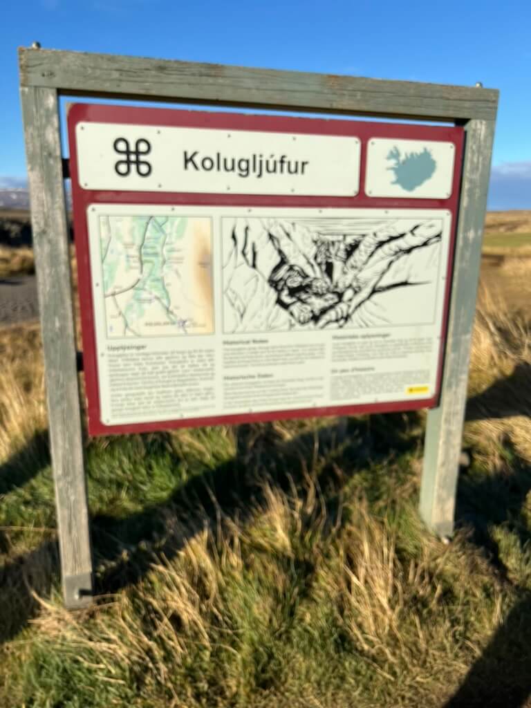 A wooden signboard titled "Kolugljúfur" stands on grassy terrain under a clear blue sky. It features a map, text, and an illustration, conveying an informational tone.
