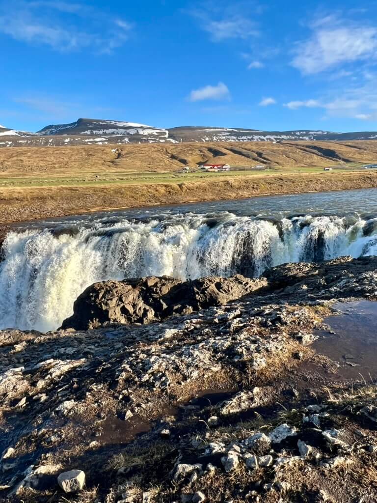A cascading waterfall flows over rocky cliffs, with a clear blue sky and snow-dusted mountains in the background, creating a serene and picturesque scene.