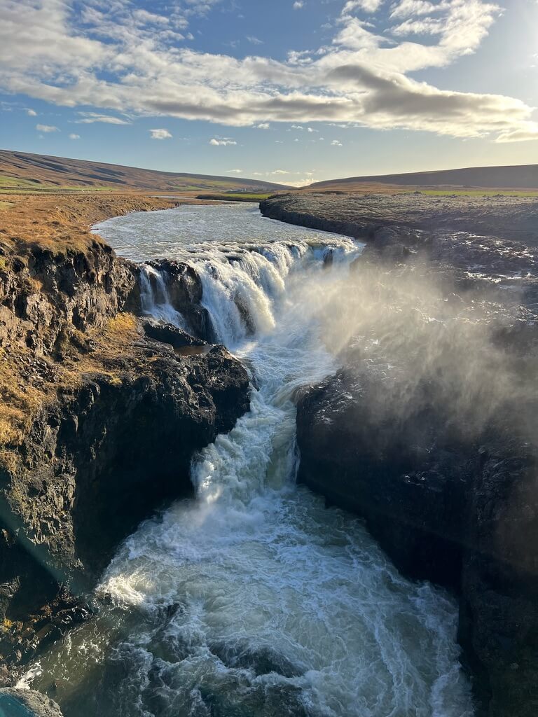 A scenic waterfall flows over rocky cliffs into a river under a bright, partly cloudy sky. The sunlight and mist create a serene, powerful atmosphere.