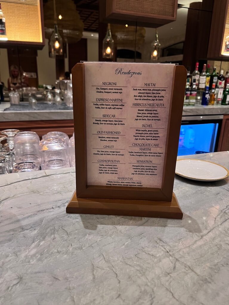 A cocktail menu titled "Rendezvous" sits on a marble bar counter. The bar is well-lit, with various liquor bottles in the background, creating a cozy, elegant atmosphere.