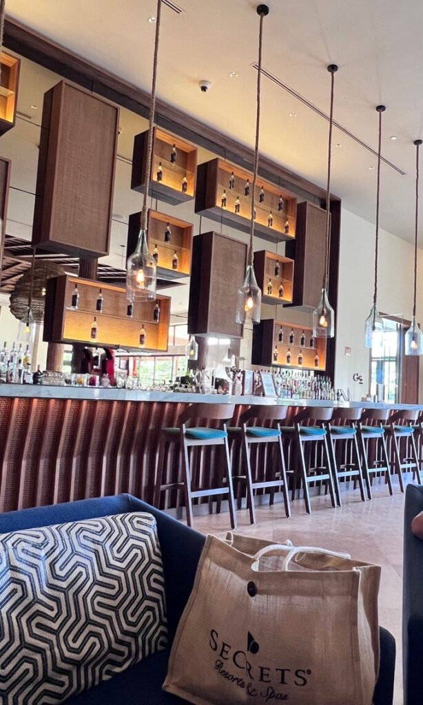 Modern bar interior with hanging lights and wooden shelves displaying bottles. A plush sofa with a geometric cushion sits in the foreground, next to a bag labeled "Secrets Resorts & Spa." The atmosphere feels stylish and inviting