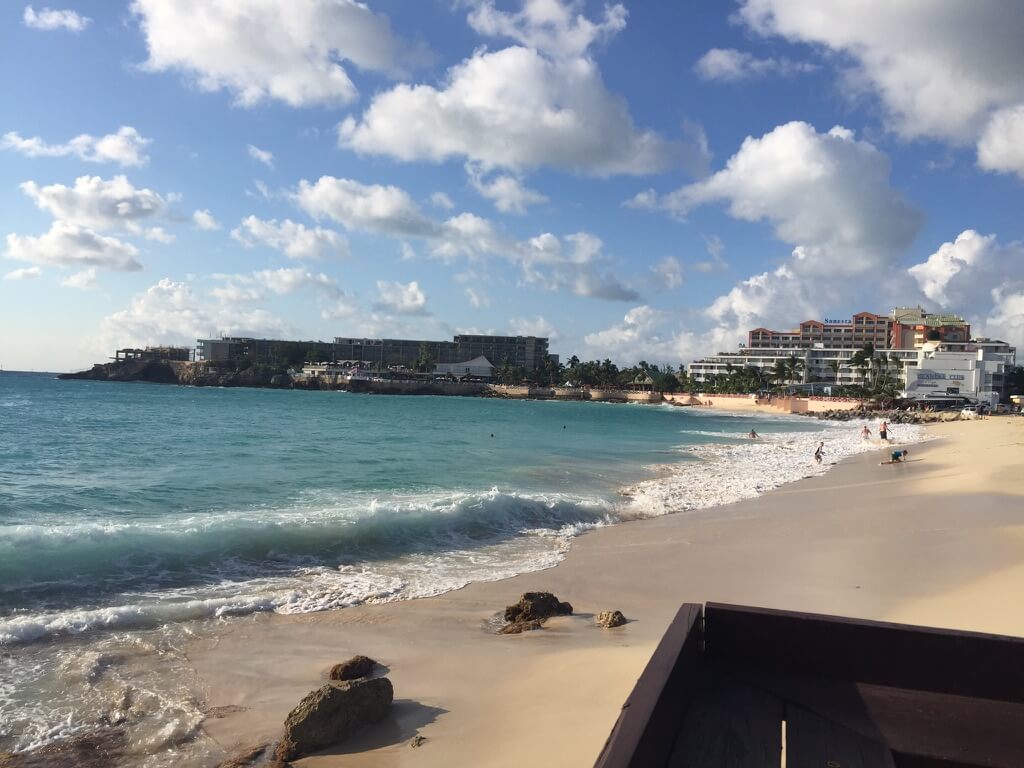 Sunny beach scene with turquoise waves gently crashing onto the sandy shore. People are enjoying the water. Hotels line the distant coastline.