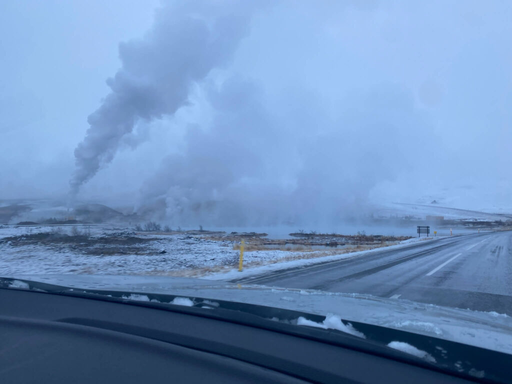 Snowy landscape with a road curving into the distance, steam rising from multiple geothermal vents on the left. Overcast sky, cold and misty atmosphere.