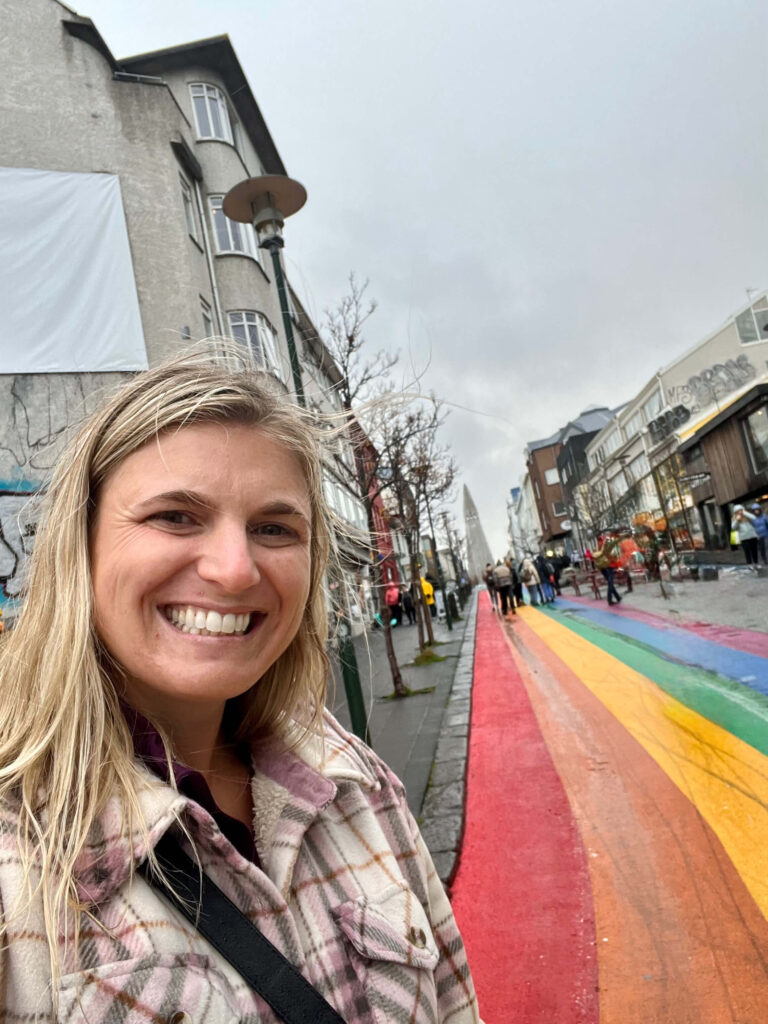 A woman smiles in front of a rainbow-painted street in an urban setting. The sky is overcast, creating a vibrant contrast with the street's colors.