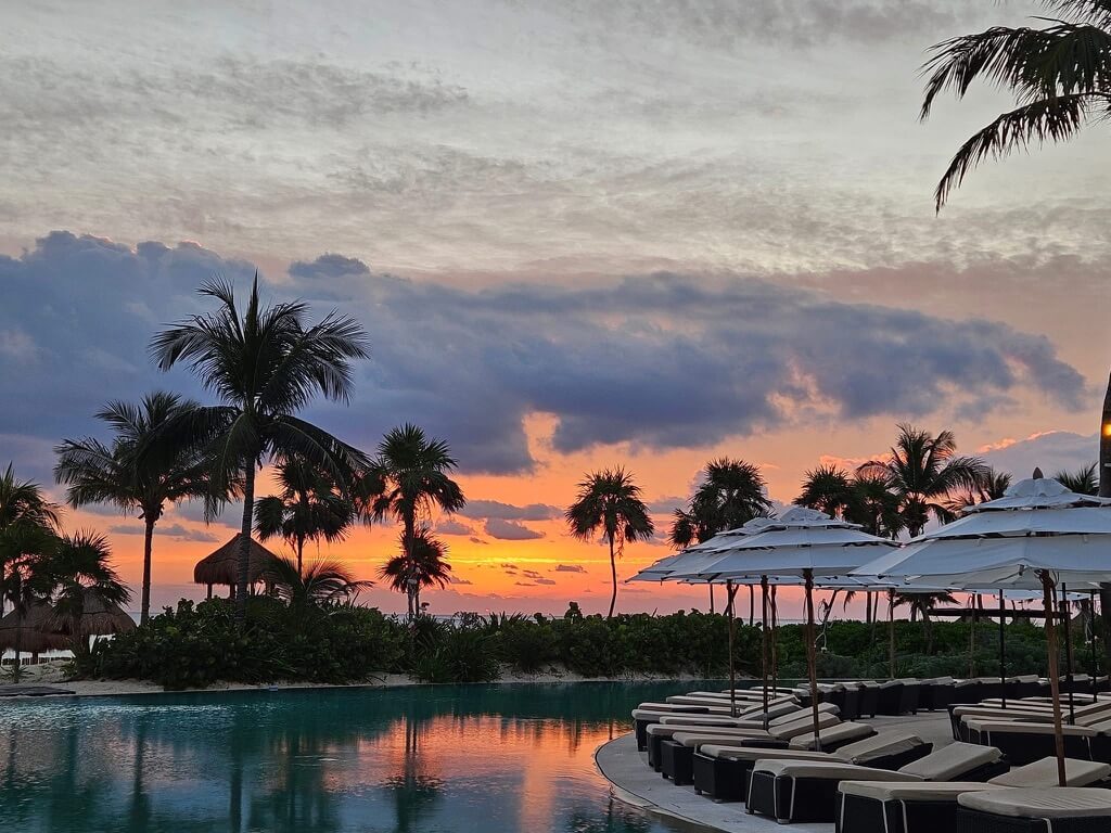 Tranquil sunset over a tropical resort, with silhouetted palm trees and cozy loungers under umbrellas by a reflective pool, evoking relaxation.