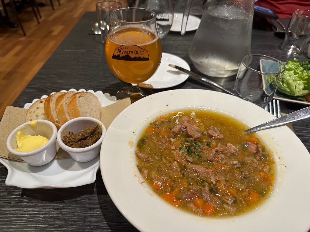 A bowl of hearty soup with meat and vegetables, served with sliced bread, butter, and relish. A glass of amber beer and a water jug are nearby on a dark table.