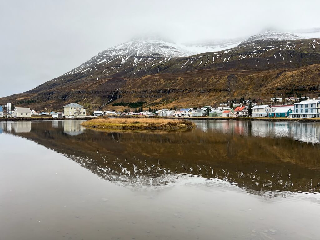 Snow-capped mountains overlook a serene lake reflecting pastel-colored houses in a quaint village, under a cloudy sky, evoking tranquility.