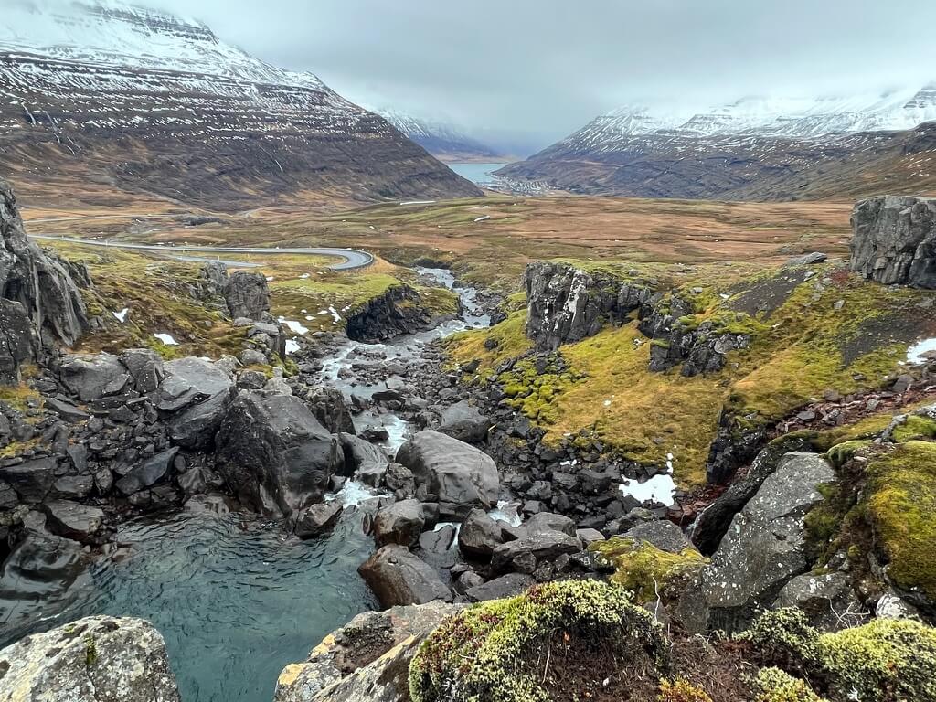 Mountain landscape with rugged cliffs and a river flowing through green mossy terrain. Snow-capped peaks in the background under a cloudy sky. Peaceful and majestic.