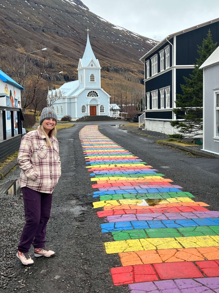 A woman in a plaid jacket stands smiling next to a vibrant rainbow-colored cobblestone path leading to a light blue church against a mountain backdrop.