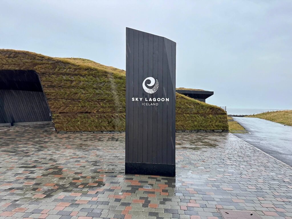 A rainy day scene at Sky Lagoon, Iceland. A black sign stands on a wet, patterned walkway, leading to a grassy mound under an overcast sky. Tranquil mood.