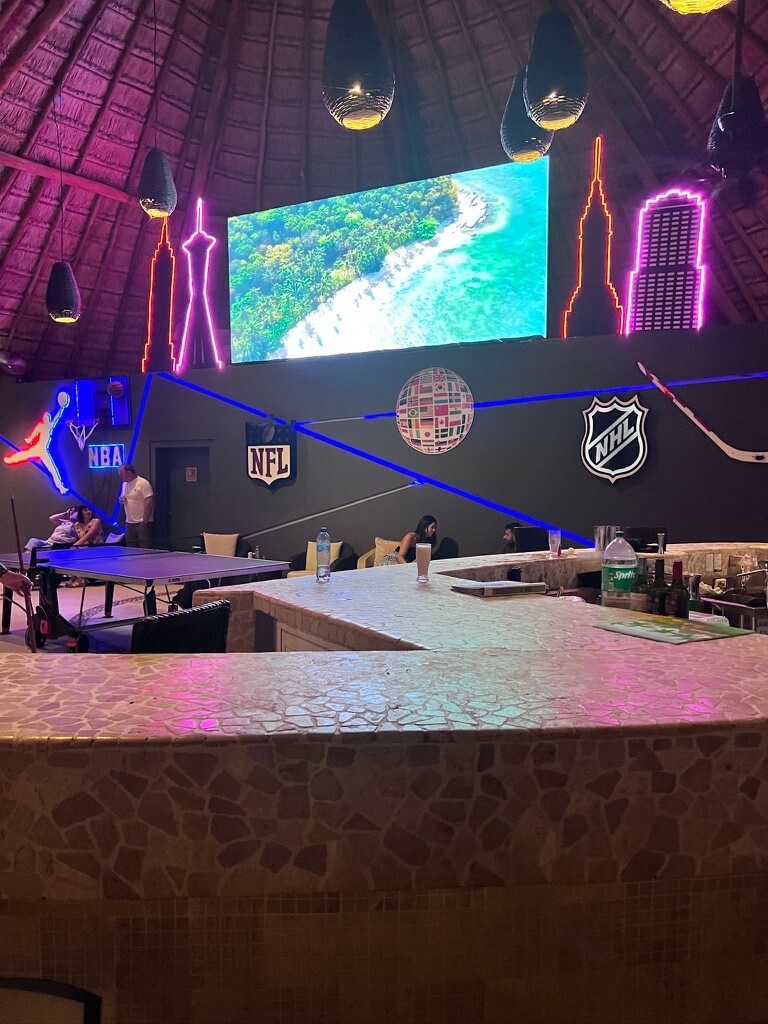 Tropical-themed bar with mosaic counter, neon sports logos, and a large screen showing a beach. People playing ping pong, creating a lively atmosphere.