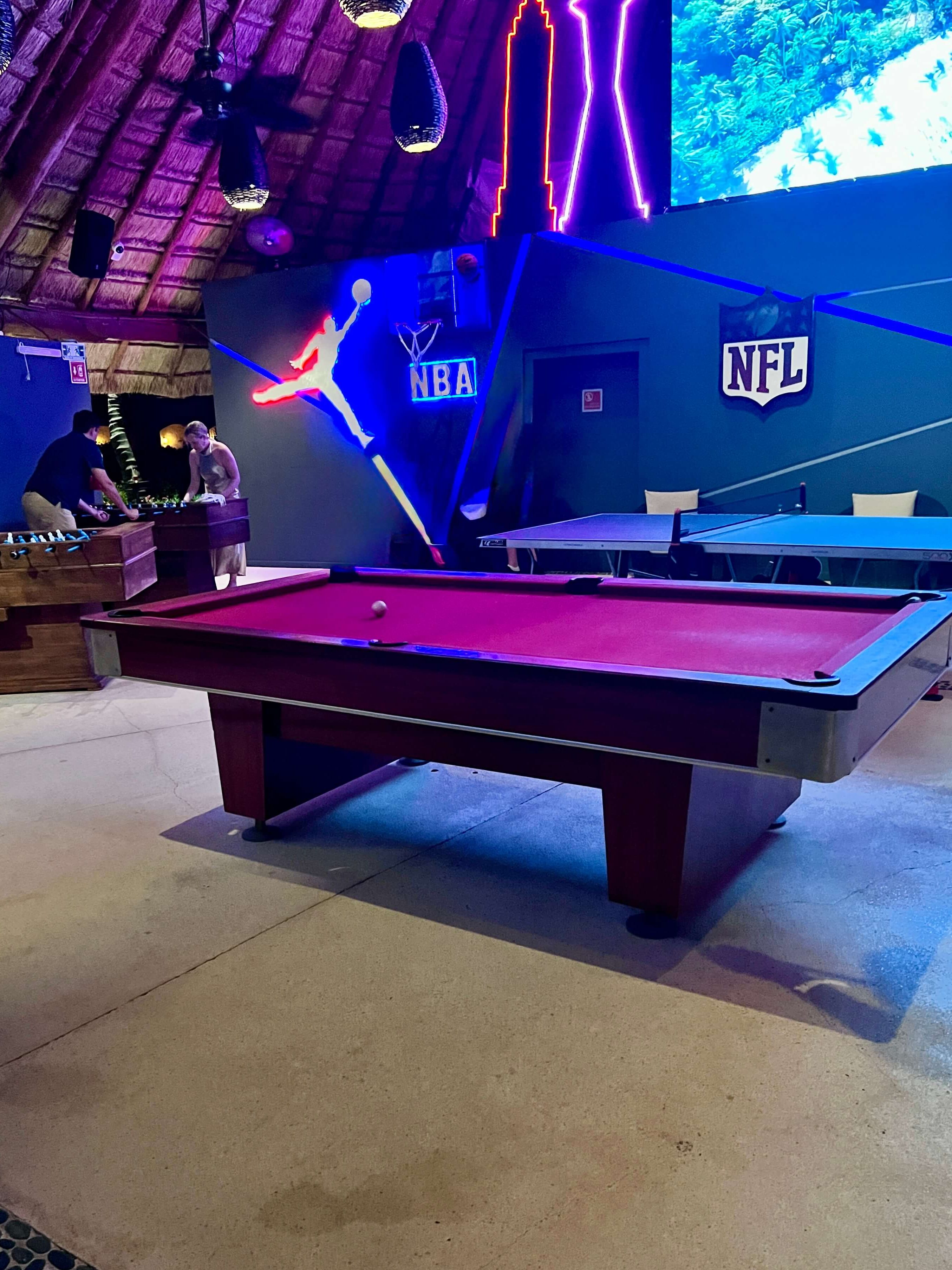 A game room with a red pool table in the foreground, foosball table on the left. Neon NBA and NFL signs adorn the walls, creating a vibrant ambiance.