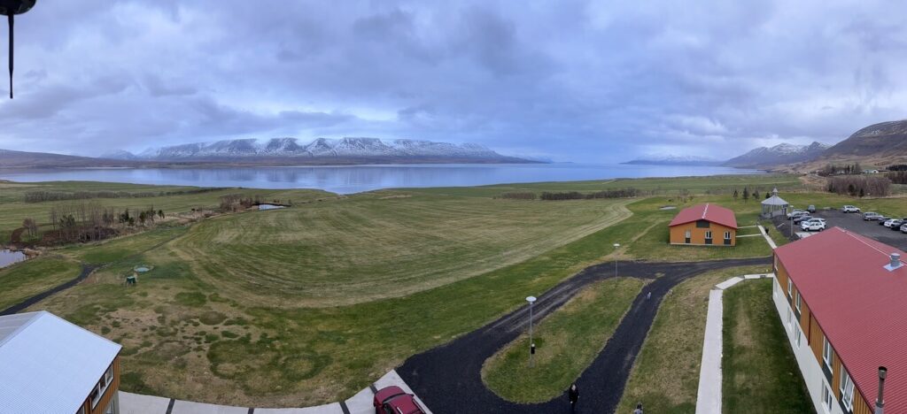 Expansive view of a grassy field leading to a calm lake, framed by snow-capped mountains under a cloudy sky. Buildings with red roofs add contrast.