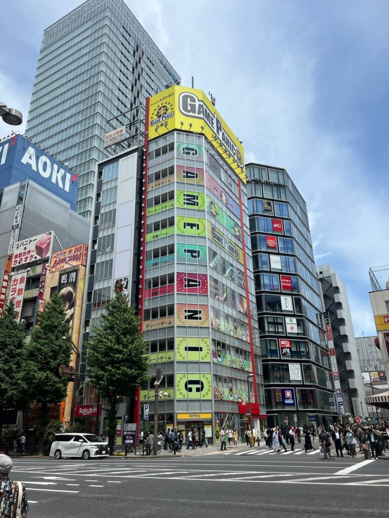 Busy street scene in Japan featuring colorful high-rise buildings with bright signage. People cross at the intersection under a clear blue sky.