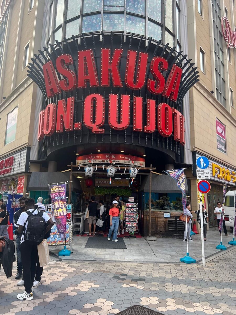 Front view of a bustling Don Quijote store in Asakusa, Tokyo. Large red signage dominates above the entrance, with people entering and exiting. Energetic atmosphere.