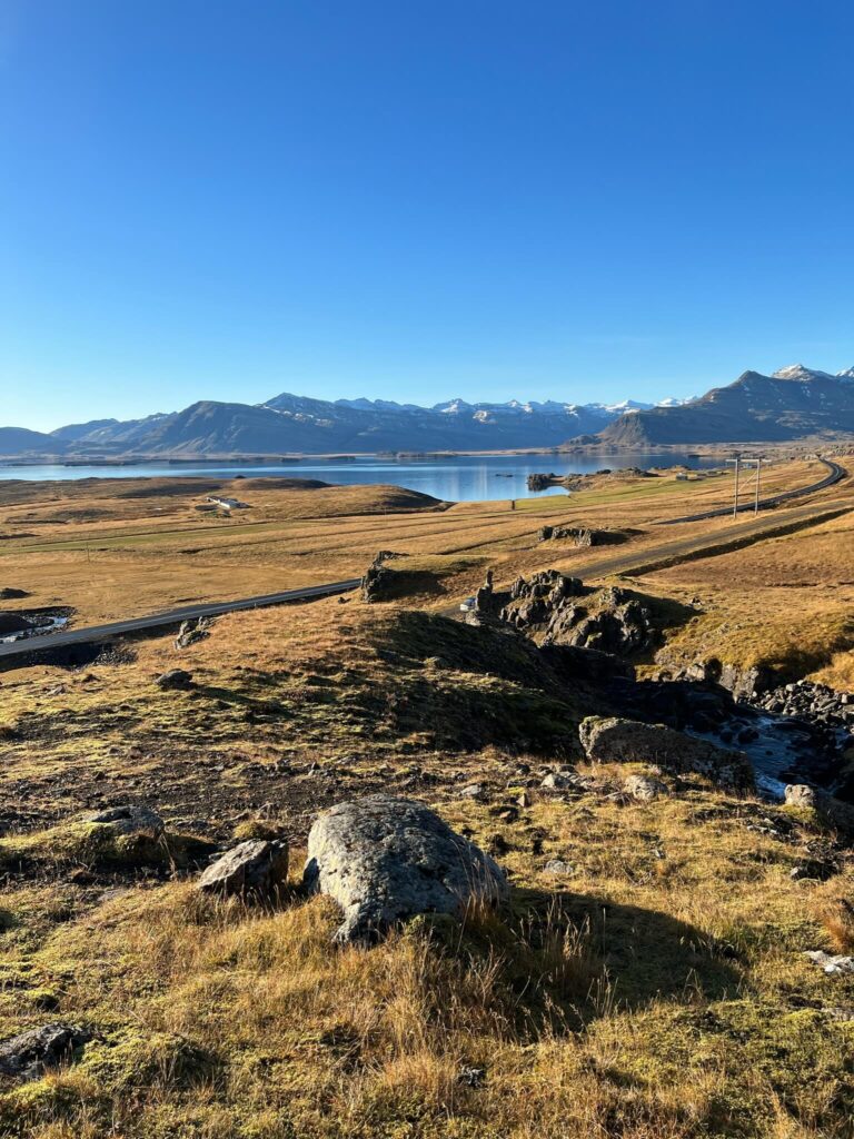 Expansive landscape showing golden fields, rocky terrain, and a serene lake under a clear blue sky. Snow-capped mountains line the horizon, evoking tranquility.