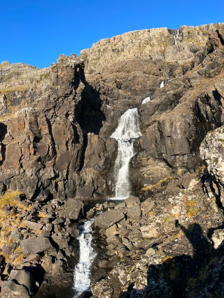 Rocky waterfall cascading down a rugged cliff under a clear blue sky. Sunlit rocks and scattered vegetation add texture and contrast. Shadow visible in foreground.