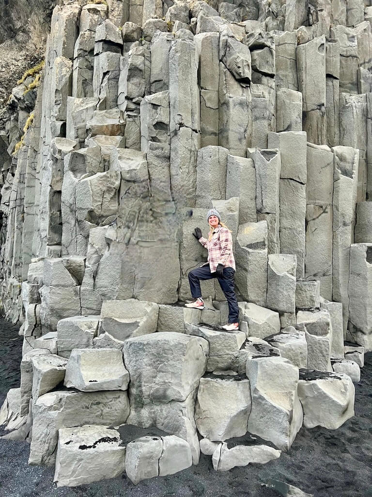 A woman in a plaid jacket and beanie poses on geometric basalt columns. The scene conveys a sense of adventure and wonder at nature’s formations.