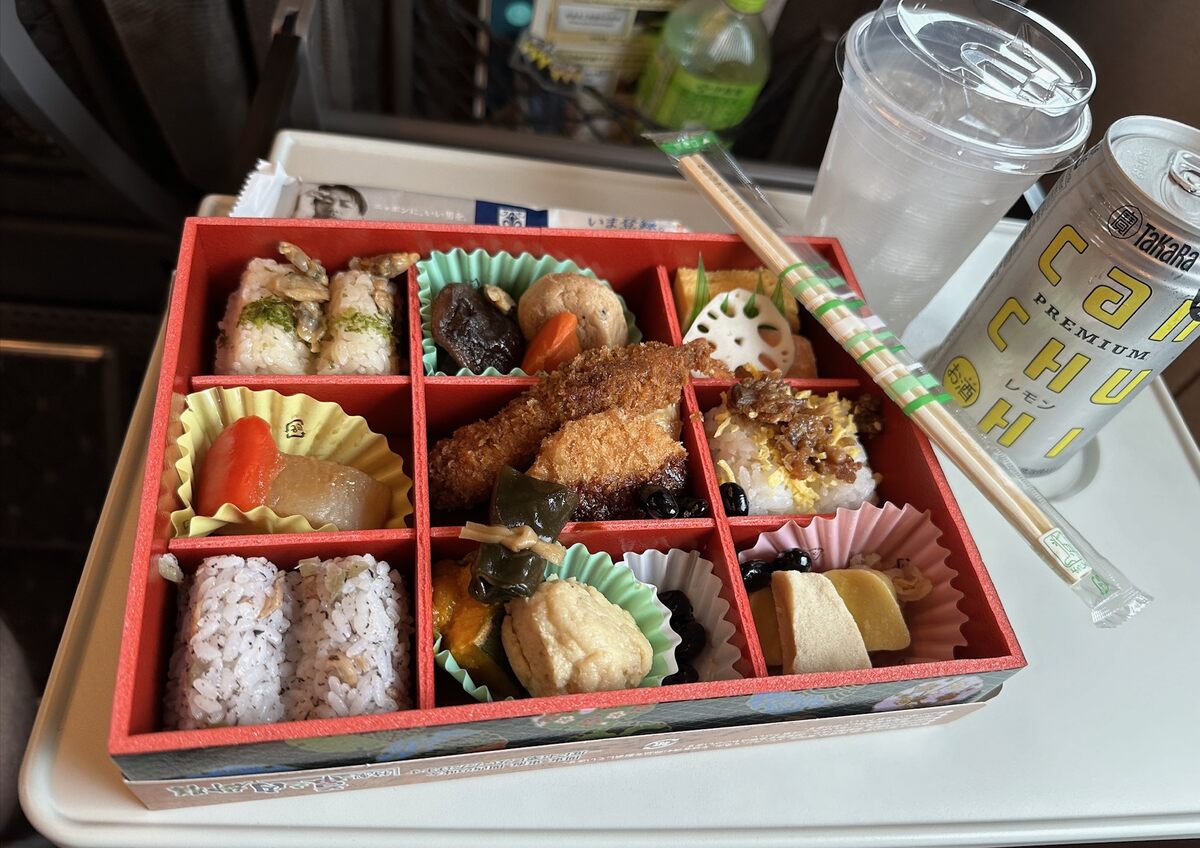 A colorful bento box with nine compartments featuring various Japanese dishes like sushi, tempura, and vegetables. Accompanied by chopsticks, a drink can, and a plastic cup on a tray.