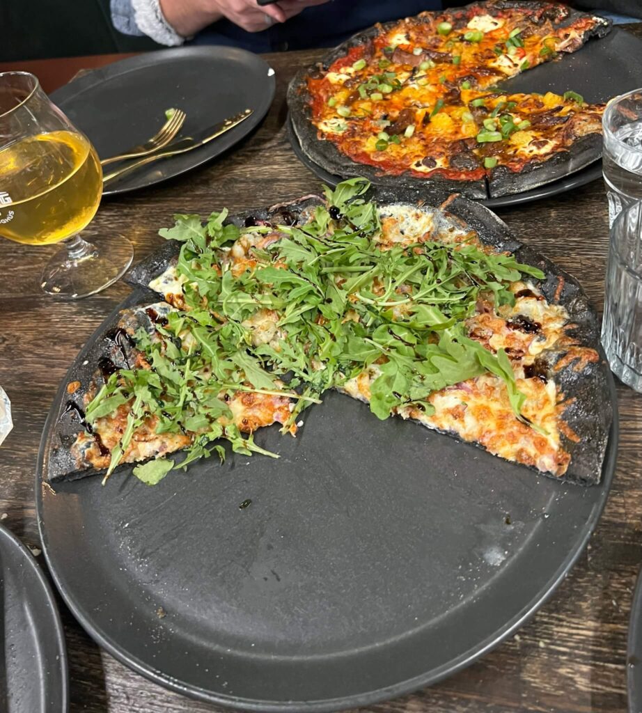 Charcoal-crust pizzas on dark plates; one topped with arugula and olives, the other with cheese and vegetables. A glass of beer and utensils nearby.