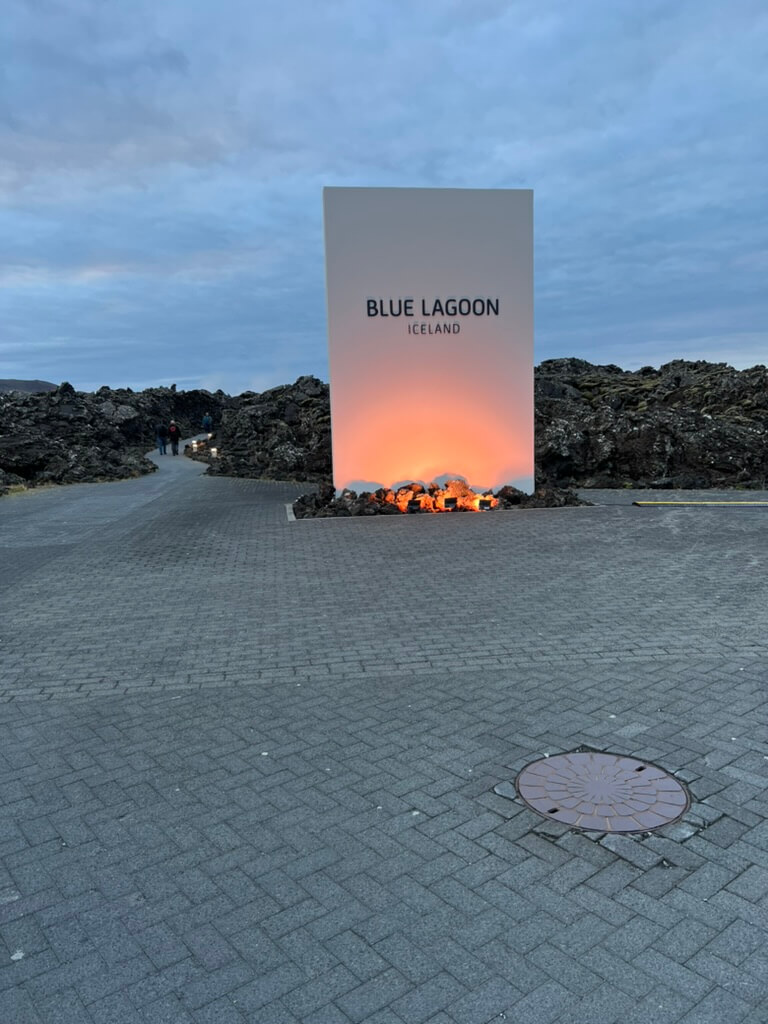Large sign reading "Blue Lagoon Iceland" stands against a backdrop of rocky landscape. Warm light illuminates the base, creating a serene atmosphere.