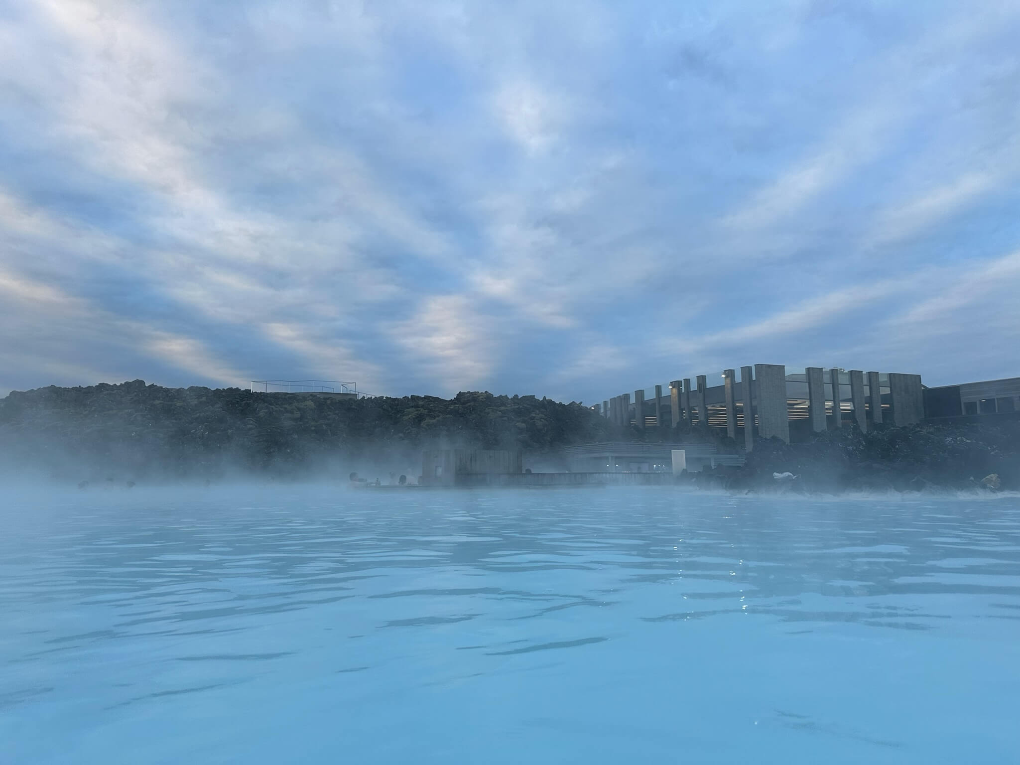 Geothermal lagoon with steaming blue water under a cloudy sky. Modern buildings and rocky landscape in the background. Calm and serene atmosphere.