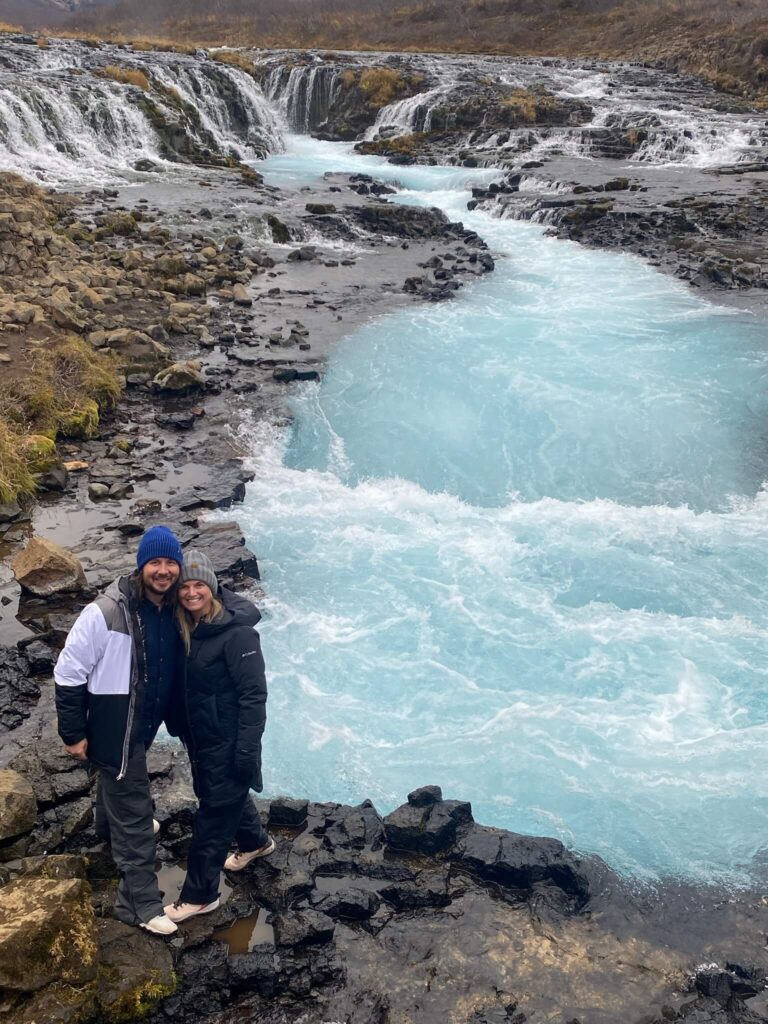 Couple in winter clothing smiles by a bright blue, cascading waterfall surrounded by rugged rocks and sparse vegetation, conveying adventure and joy.