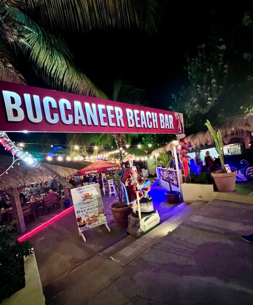 A lively beach bar entrance with a bright red sign reading "Buccaneer Beach Bar." Nighttime fairy lights and palm trees create a tropical, festive vibe.