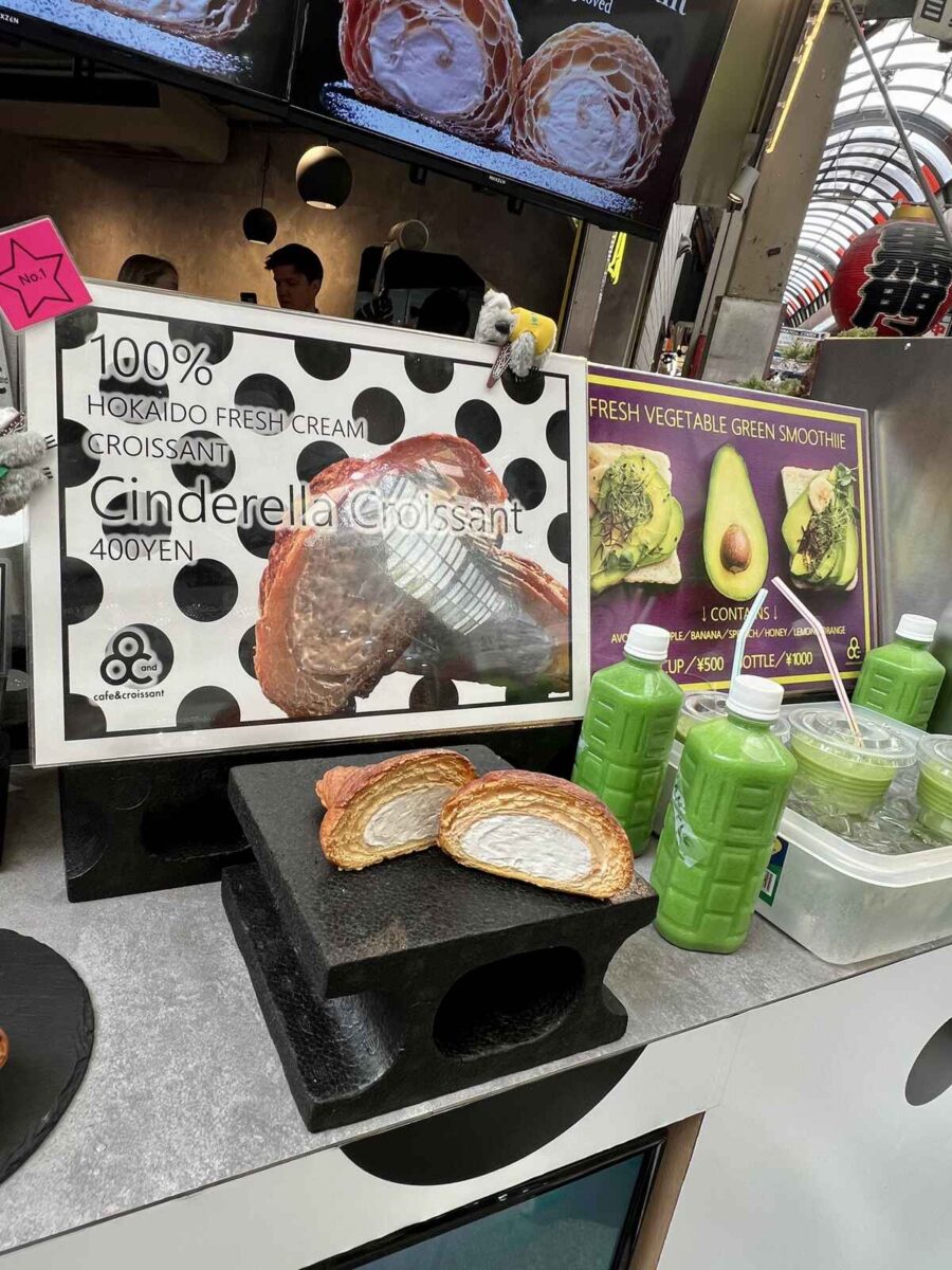 A display at a food stall showcases a promotional sign for "Cinderella Croissant" with a cross-section image revealing its creamy filling. Two halves of the croissant sit on a black stand. Nearby, green smoothies are lined up in bottles and cups. The setting conveys a vibrant, enticing atmosphere.