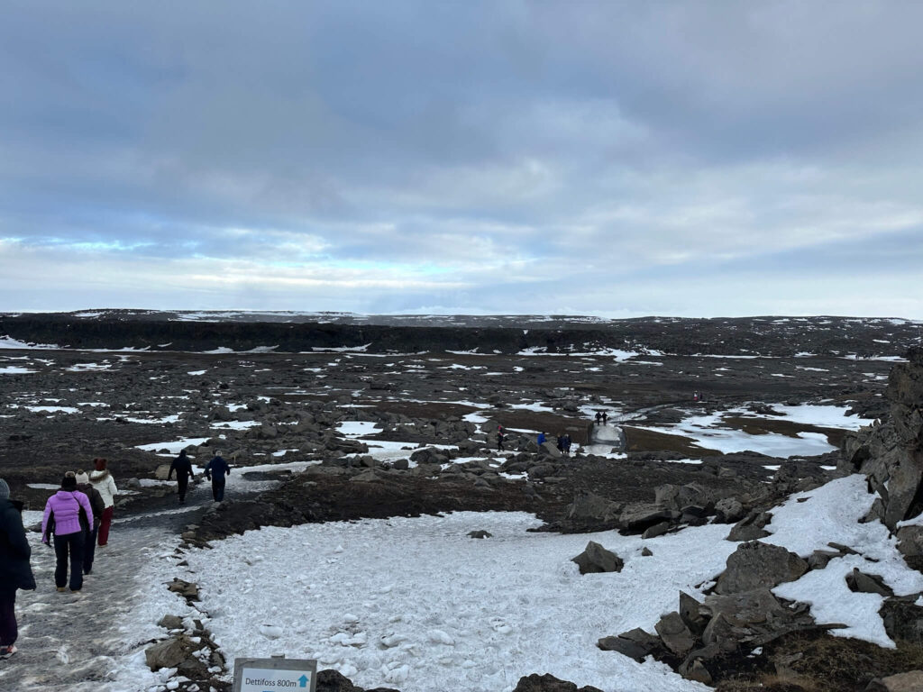 Snow-covered rocky landscape leading to Dettifoss waterfall, under a cloudy sky. Small groups of people walk along the path, creating a serene, adventurous tone.