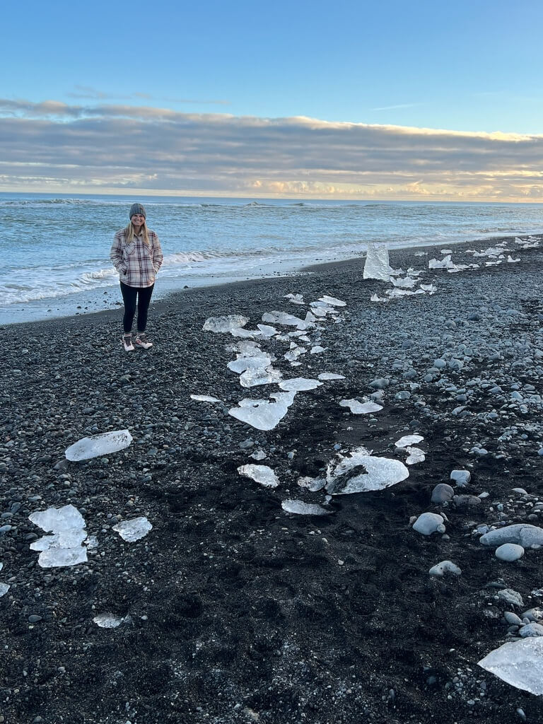 A woman stands on a pebbled beach scattered with ice chunks under a calm, partly cloudy sky. The ocean stretches to the horizon, conveying a serene atmosphere.